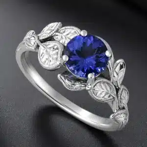 White gold engagement ring with a vibrant round tanzanite, engraved leaf accents, milgrain details, and a floral-inspired design.