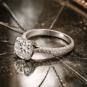 Vintage hand engraved solitaire engagement ring with round brilliant diamond and milgrain detailing, resting on a cracked antique mirror, Art Deco inspired heirloom design.