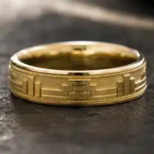 Men's 6mm Art Deco wedding band with geometric architectural pattern, keystone center design, textured background, and polished gold finish.